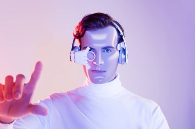 Cyborg in digital eye lens and headphones pointing with finger on blurred foreground on purple background clipart