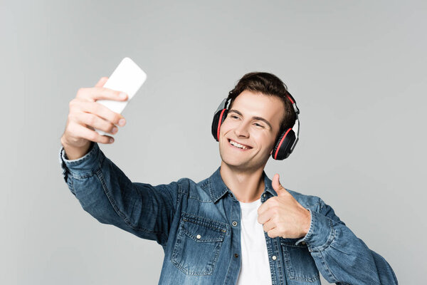 Cheerful man in headphones showing like while taking selfie with smartphone isolated on grey