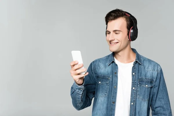 Radosny Mężczyzna Dżinsowej Kurtce Korzystający Smartfona Słuchawek Odizolowanych Szaro — Zdjęcie stockowe