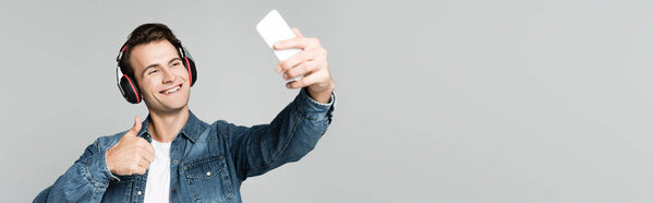 Smiling man showing like while taking selfie with smartphone and using headphones isolated on grey, banner