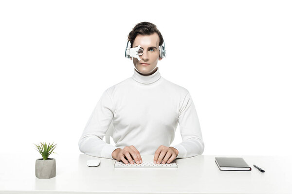 Cyborg in headphones and eye lens using computer keyboard near plant and notebook isolated on white