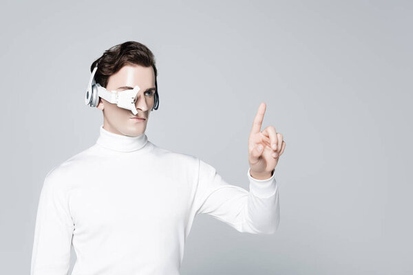 Cyborg in headphones and eye lens pointing with finger isolated on grey