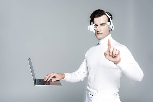 Киборг в наушниках трогает что-то рядом с ноутбуком в воздухе, изолированном на сером