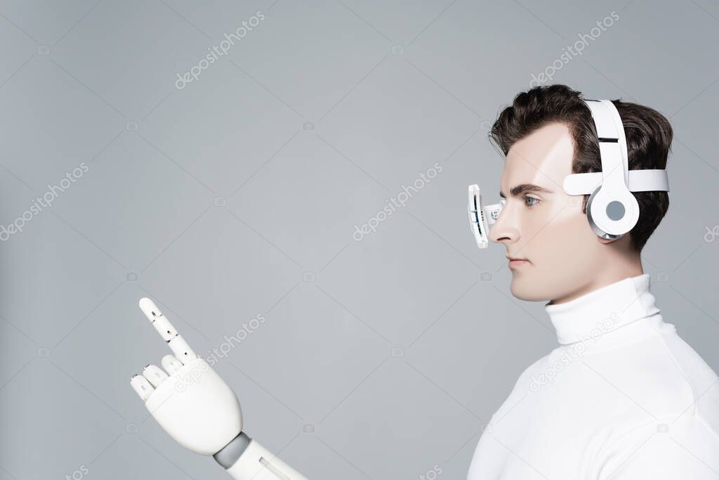 Brunette cyborg in eye lens, headphones and artificial hand pointing with finger isolated on grey