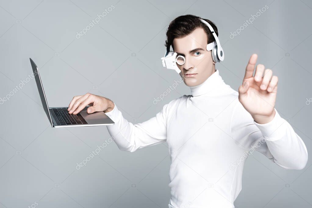 Cyborg man in headphones touching something while using laptop in air isolated on grey