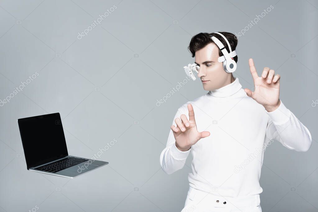 Cyborg in headphones touching something and looking at laptop with blank screen in air isolated on grey