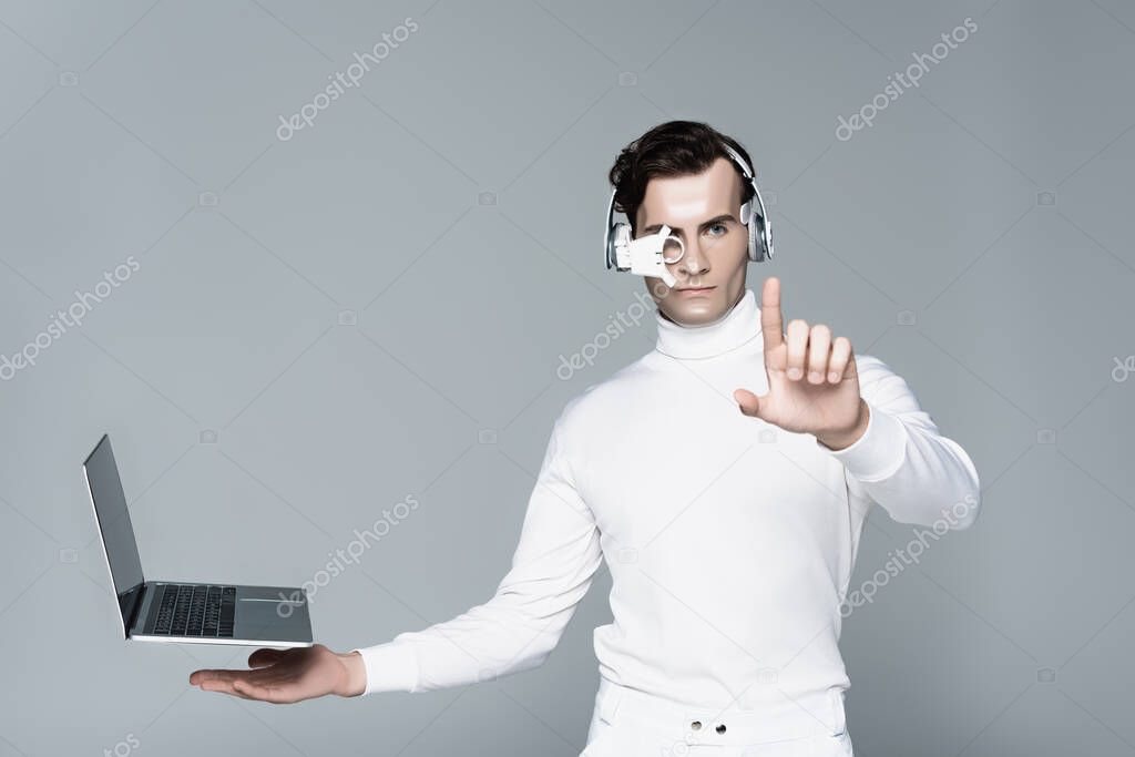 Cyborg in headphones touching something near laptop with blank screen levitating in air isolated on grey