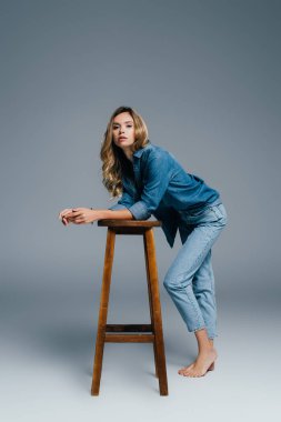 sensual woman in denim shirt and jeans leaning on high stool on grey clipart