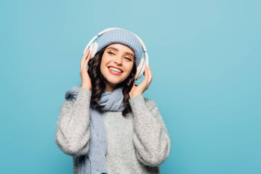Kış kıyafetli mutlu esmer kadın mavi kulaklıkla müzik dinlerken başka tarafa bakıyor.