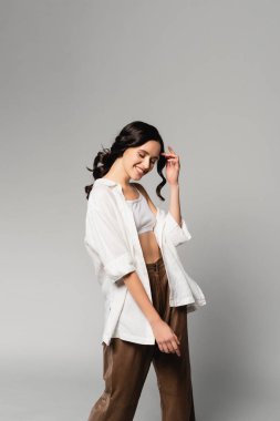 Gülümseyen esmer, gözleri kapalı, günlük kıyafetler giyen ve gri üzerinde poz veren bir kadın.