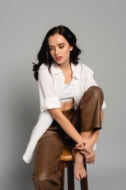 Kıvırcık esmer kadın gri taburede poz verirken gözlerini kaçırıyor.