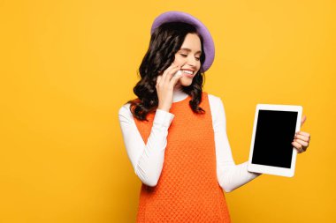 Sarıda izole edilmiş dijital tablete bakarken bereli, gülümseyen esmer kadın cep telefonuyla konuşuyor.