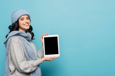 Kışlık kıyafetli neşeli esmer kadın mavi üzerinde boş ekranlı dijital tablet gösteriyor.