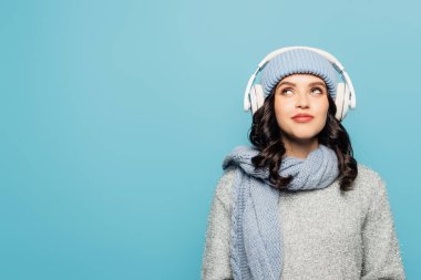Dreamy woman in winter outfit with headphones thinking while looking up isolated on blue clipart