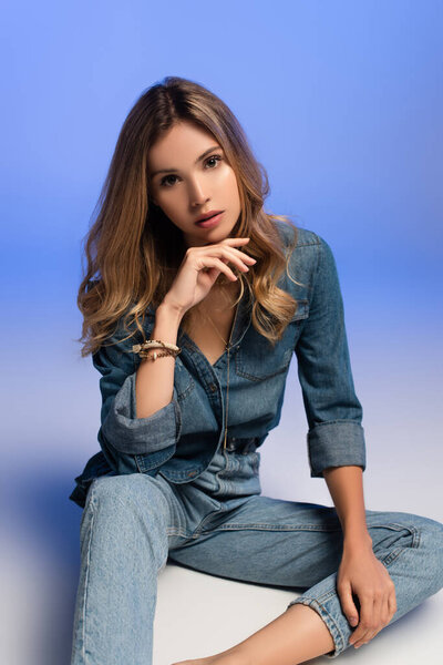 seductive young woman in denim clothes holding hand near face while posing on blue