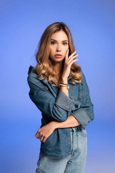 young, sensual woman in denim clothes holding hand near face isolated on blue