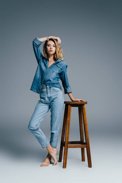 stylish barefoot woman in denim shirt and jeans leaning on high stool on grey