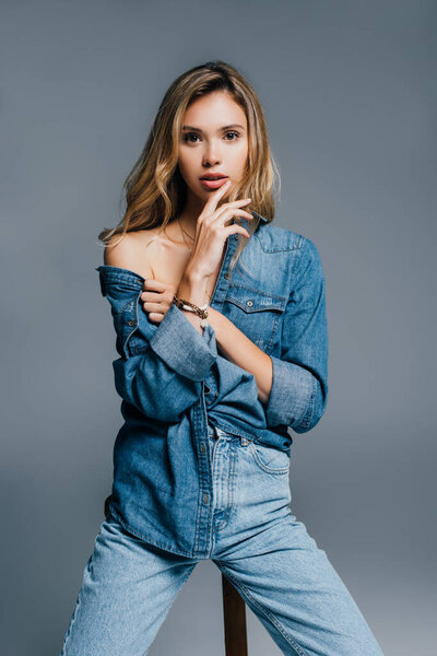 sexy young woman in denim shirt touching lips while posing with naked shoulder isolated on grey