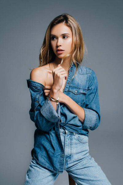 sexy young woman in denim clothes, with naked shoulder, touching hair isolated on grey