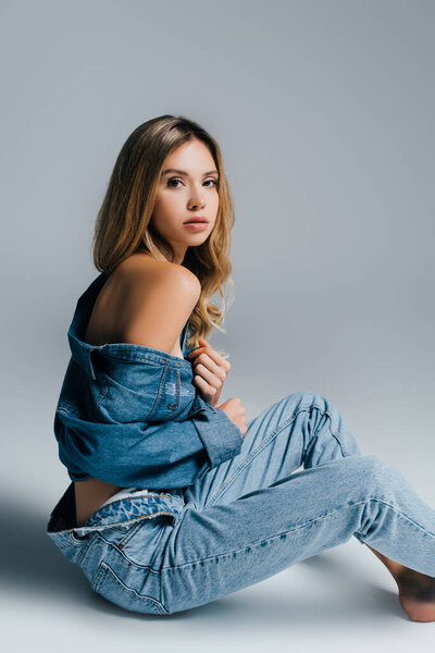 sexy young woman in unbuttoned denim shirt and jeans posing with naked shoulder on grey