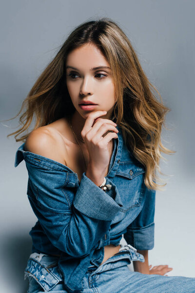 seductive young woman in unbuttoned jeans, with naked shoulder, touching chin on grey