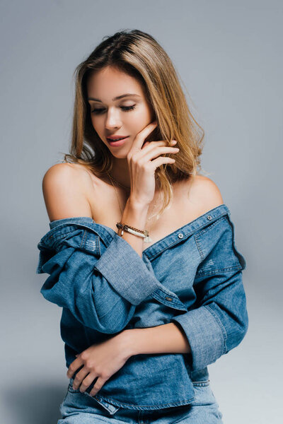 seductive young woman in unbuttoned denim shirt, with naked shoulders, touching face  on grey