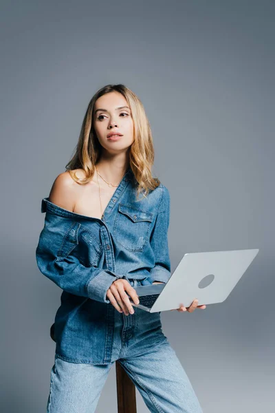 Stylowa Kobieta Dżinsowej Koszuli Nagim Ramieniem Odwracając Wzrok Trzymając Laptopa — Zdjęcie stockowe