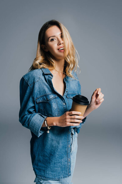 smiling woman in denim short holding paper cup while looking at camera on grey