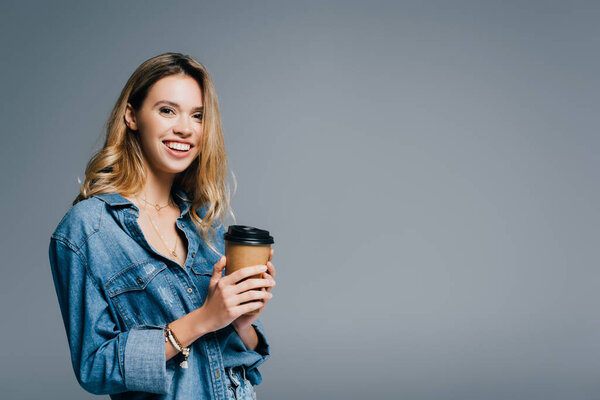 cheerful woman in denim shirt holding coffee to go and smiling at camera isolated on grey