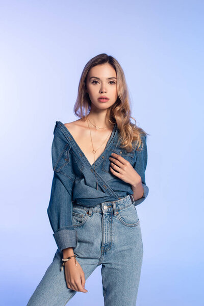 charming young woman in denim shirt and jeans looking at camera while standing isolated on blue