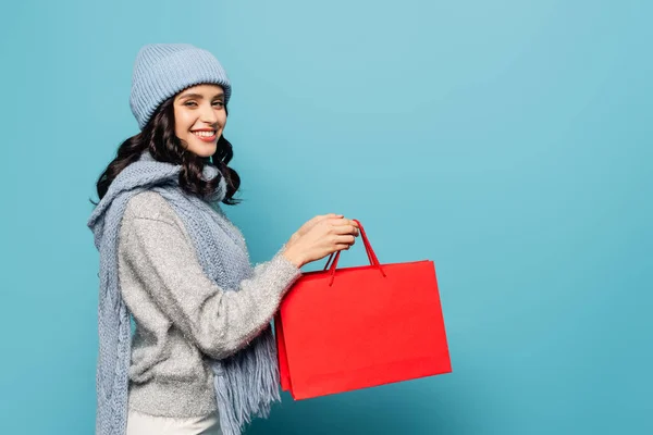 Happy brunette woman in winter outfit looking at camera while holding red shopping bags isolated on blue