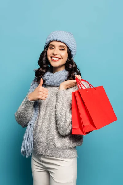 Cheerful brunette woman in winter outfit holding shopping bags and showing thumb up while looking at camera isolated on blue