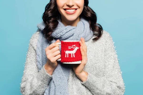 Cropped view of brunette woman in scarf and pullover holding cup with knitted holder isolated on blue