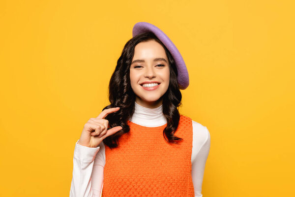 Smiling brunette woman in beret showing small amount gesture while looking at camera isolated on yellow