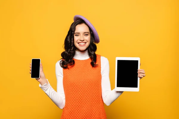 カメラを見ながらベレー帽で陽気女性は黄色に隔離されたスマートフォンやデジタルタブレットを示しています — ストック写真