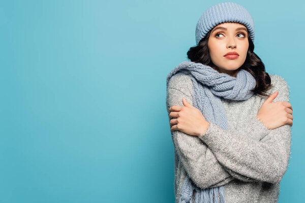 Brunette woman in winter outfit hugging herself while looking away isolated on blue