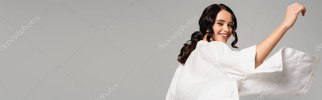 Cheerful brunette woman in white shirt looking at camera while turning around isolated on grey, banner