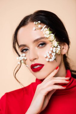 Kırmızı dudaklı ve yüzünde çiçekler olan genç bir kadın pembe kameraya bakıyor. 