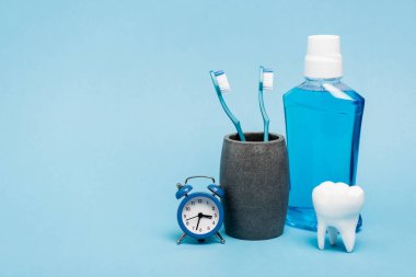 Small clock near toothbrushes, mouthwash and tooth model on blue background clipart