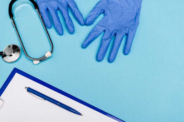 Top view of latex gloves near stethoscope and clipboard with pen on blue background