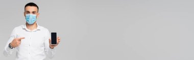 Tıbbi maskeli İspanyol bir yönetici gri pankartta boş ekranlı akıllı telefonu işaret ediyor.
