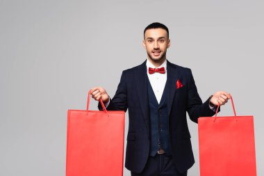 happy hispanic man in elegant suit holding red shopping bags isolated on grey clipart