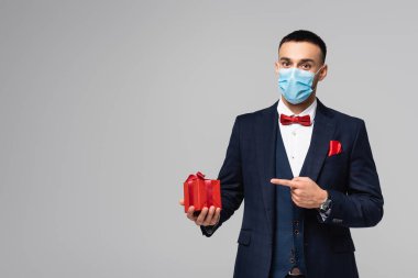 young hispanic man in elegant suit and medical mask pointing at red gift box isolated on grey clipart