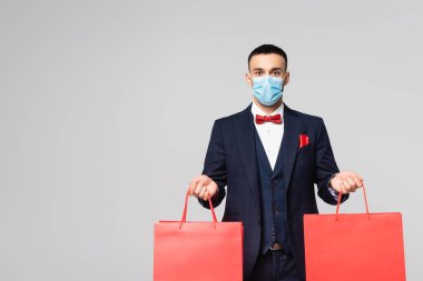 elegant hispanic man in medical mask holding red shopping bags isolated on grey clipart