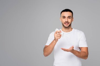Beyaz tişörtlü gülümseyen İspanyol adam gri üzerine izole edilmiş bir el kremi tüpü tutuyor.
