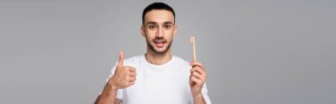 Neşeli İspanyol adam baş parmağını gösterip diş fırçasını gri pankartta tek başına tutuyor.