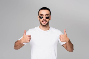 Göz bantlı İspanyol adam gri kameraya gülümserken baş parmağını kaldırıyor.