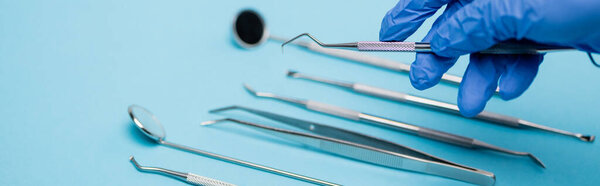 Cropped view of dentist in latex glove holding tool near dental set blurred on blue background, banner 