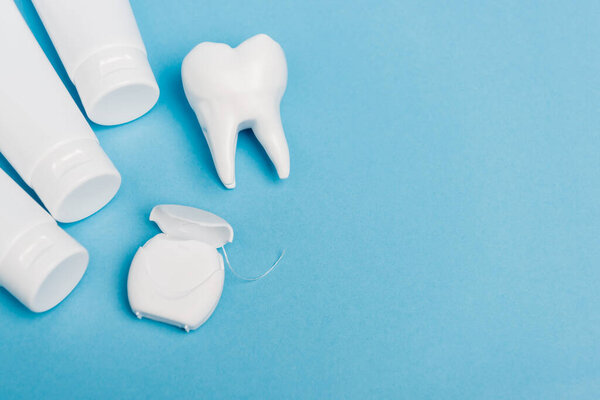 Top view of dental floss near tooth model and tubes with toothpaste on blue background