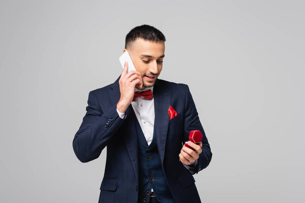 smiling hispanic man in blue suit talking on smartphone while holding jewelry box isolated on grey